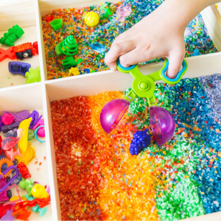 childs hand dipping into a box of multicoloured sand
